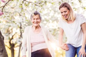 home care in Laughlin, NV (1) young woman in white holding hands with a smiling senior woman outdoors on a sunny day, surrounded by greenery.