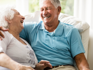 A couple sit on a couch laughing, with husbands arm around wife. Home care in Mesquite, NV