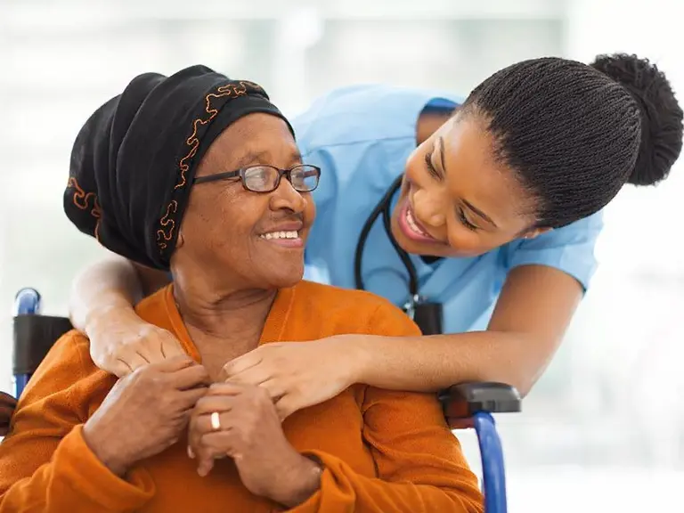Smiling caregiver in blue scrubs leaning warmly toward a senior woman in a wheelchair wearing an orange sweater and black headscarf, reflecting compassionate in-home senior care