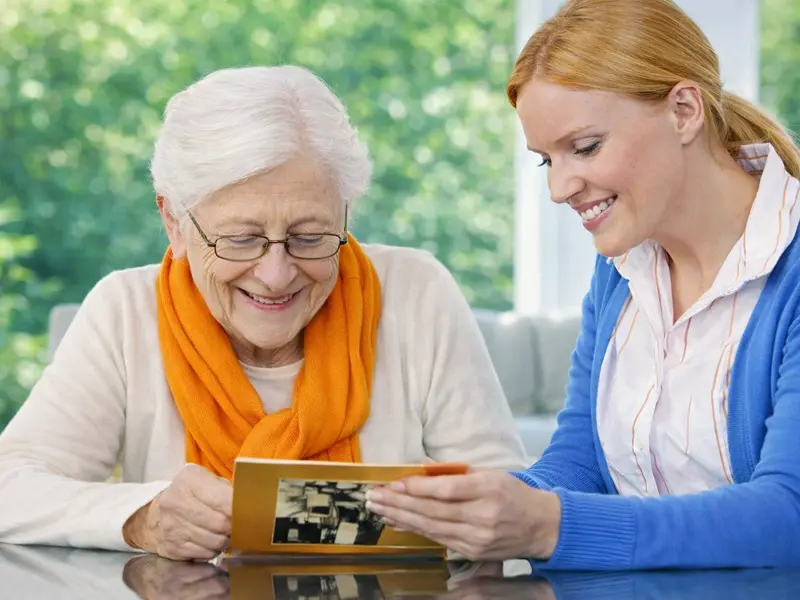 Smiling senior woman wearing an orange scarf looking at cherished photographs with a caregiver in a blue cardigan, reflecting supportive dementia care in a warm home environment.
